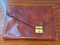 Front view of cognac leather portfolio showing envelope flap with gold-toned lock hardware and visible surface wear.