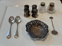 Full lot view showing two enamel badge spoons, Dutch coin spoon, four metal salt shakers, two glass salt shakers, and a decorative silver dish.