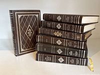 Side view showing one book standing up and six stacked horizontally, displaying decorative covers and gold embossed spine details.