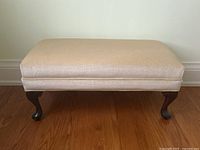 Front view of rectangular upholstered footstool with dark wood curved legs on hardwood floor against plain wall.