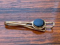 Side view of the gold tie clip featuring an oval black onyx stone, showing the sleek intertwined gold design and polished stone surface.