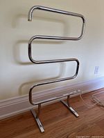 Front view of the chrome metal towel warmer showing the S-shaped zig-zag heated bars and the two-legged standing base, power cord and switch.