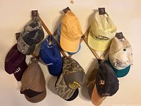 Photo showing 10 assorted baseball caps hanging on a wooden rack in various colors and patterns including camo, yellow, blue, tan, black, and cream with logos and text.