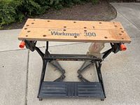 Front angled view of Black & Decker Workmate 300 workbench showing wooden top, black metal frame, and orange handles at sides.