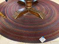 Round braided rug shown with base of wooden furniture for scale. Rug has a concentric circle braided pattern with red, blue, tan, and maroon colors. Rug surface shows debris.