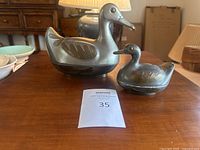 Both duck and duckling trinket boxes displayed on a wooden table with lamp and other decor in background, showing size difference and brass feather details