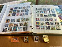 Two open binders with multiple collectible and used stamps, arranged in rows and columns on album pages. Four collectible pins placed in front of the binders.