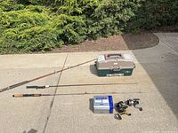 Fishing poles, reels, and tackle box placed on concrete surface with greenery background.