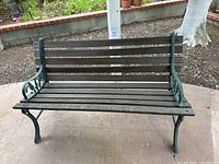 Front view of outdoor bench with wrought iron frame and wooden slats seat and backrest on concrete patio with garden in background.