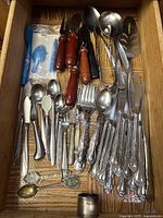 Top-down view of a kitchen drawer with assorted silverware including forks, spoons, knives, and cheese cutters with wooden handles.