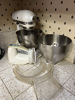 White KitchenAid Classic stand mixer with wire whisk attachment, two stainless steel mixing bowls placed on tiled countertop, and white Hamilton Beach hand mixer with power cord coiled on handle.