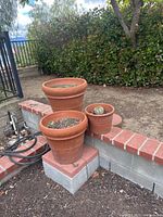 Three terracotta planters resting on brick and concrete blocks in an outdoor garden area, surrounded by soil and plants.