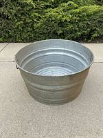 Silver galvanized metal tub placed on concrete with green shrub background, showing interior and ribbed bottom.