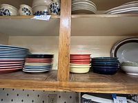 Shelves displaying multi-colored Fiesta dinner and salad plates stacked in piles by color including pastel blue, and pink, with additional stacks of Fiesta bowls in red, yellow, blue and white as well as white Williams-Sonoma nesting bowls.