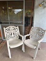 Pair of beige plastic patio chairs side by side showing ornate floral and scrollwork design on backs, armrests, and seats.