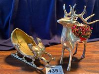 Reindeer candle holder with red berry garland and brass sleigh side by side on wooden surface with dark blue background.