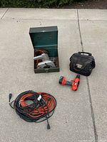 Photo of Black and Decker circular saw inside a green metal case, cordless drill, soft carrying case, and extension cords on ground