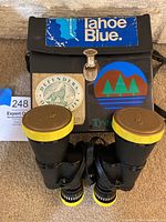Photo showing black textured binoculars with yellow caps and knobs in front of a black vinyl case with stickers