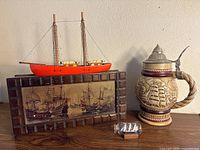 Image shows a collection of nautical-themed items including an Avon beer stein decorated with ship reliefs and a pewter lid, a wooden sailboat model in red, a ship in bottle, and a wooden decorative box with vintage sailing ship artwork.