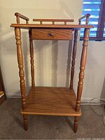 Front view of the small wooden side table showing decorative turned legs, drawer, lower shelf, and raised edges on three sides of the tabletop.