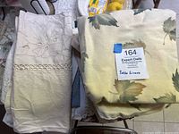 Photo showing stack of folded table linens including beige embroidered linen with floral cutwork and autumnal leaf patterned cloth.