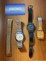 Photo of three vintage wristwatches along with Seiko certificate of guarantee on wooden surface. Watches include a Seiko with silver metal bracelet and additional gold-tone stretch bracelet, a black Pulsar with mesh strap, and a gold-tone Armitron with light textured dial.