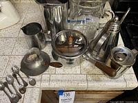 Top down view of the pitchers, pan with lid, server on saucer, and teaspoons arranged on tiled surface