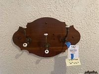 Front view of the vintage wooden wall coat rack with three brass and white ceramic hooks on a textured wall.