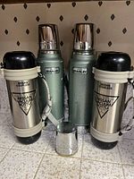 Four vintage coffee pitchers including two green Aladdin Stanley vacuum bottles and two stainless Nissan Vacuum Bottles with black plastic tops and bottoms, arranged on tiled surface.