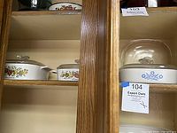Three vintage casserole dishes in a cabinet, showing fruit/vegetable and blue floral designs with glass lids