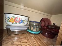 Shelf with various vintage cookware including a white enamel Corning bowl with floral pattern, clear glass storage containers with lids, and purple glass dishes.
