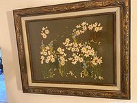 Framed vintage embroidered floral art hanging on a wall, showcasing detailed white and yellow flowers embroidered on green fabric with ornate wooden frame.