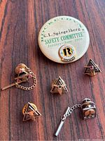 Six gold pins displayed with safety committee button background; four triangular enamel NASA pins, one square tie tack with stones and chain, one rectangular tie tack with chain.