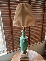 Vintage table lamp with green ceramic base featuring white decorative patterns, square ornate base, and beige pleated shade. Lamp shown unlit.