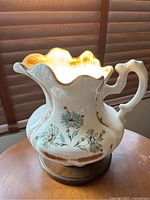 Front angle of the ceramic floral pitcher lamp showing ruffled rim and ornate handle.