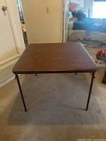 Front angled view of vintage Samsonite card table with brown vinyl top and folding legs in a carpeted room.