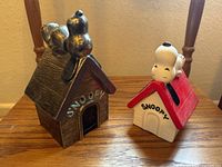 Two Snoopy dog house figurines, one metal coin bank and one ceramic house with Snoopy lying on red roof.