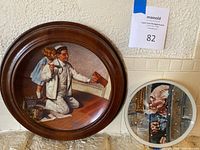 Two Norman Rockwell plates displayed: a larger framed plate with man painting and girl, and a smaller plate with man looking out a snowy door.