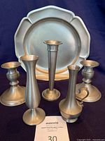 Display of all pewter items including two candle holders, three bud vases, a taller vase with handle, and a round tray in the back.
