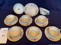 Photo showing 6 porcelain bisque tea cups on matching saucers and additional china pieces including 1 small bowl and 2 other cups with floral patterns.