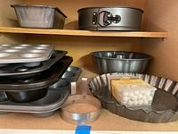 Shelf with stack of various metal baking pans including bread pan, springform cake pan, Bundt pan, fluted pie pans, and clear plastic box of ceramic pie weights.