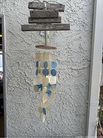 Full view of the wind chime hanging against a textured white wall showing shell pieces and the wooden sign with quotes above.