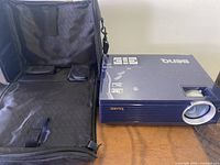 Blue BenQ digital projector with marker labels and vent grille, placed on wooden surface next to open padded carrying case.