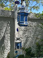 Two blue glass lanterns and a wind chime hanging outside on a vertical post against a textured wall, partially surrounded by greenery.