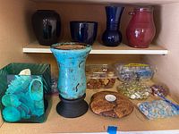 Photo showing five ceramic vases in varied colors on two shelves, burl wood tea light holder in front with decorative glass pebbles and rocks in containers and bags, green glass vase filled with seashells.