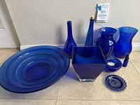 Full view of cobalt blue glassware collection showing two tall bottles, three vases, large round plate, small round plate, square vase, ring holder, and a tiny bottle with stopper.