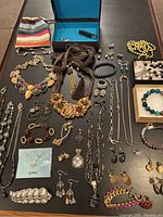 Full layout of assorted costume jewelry including necklaces, bracelets, earrings, and brooches on a black cloth with a colorful striped bag and an open jewelry box with separated smaller pieces.