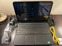 Dell Inspiron laptop open on dark wood surface, power adapter wrapped in plastic, yellow Ethernet cable coiled beside it.