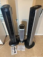 Front view of two Seville tower fans and one Lasko tower heater with remotes and manuals laid out on floor.