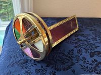 Side angled view of the brass kaleidoscope showing the round viewing end with colorful art glass pieces and the rectangular body with red textured glass and brass detailing.